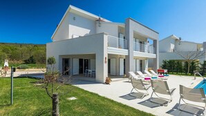 Une maison moderne à deux étages, dotée d’un balcon, d’un espace piscine avec des chaises longues et d’un petit jardin.