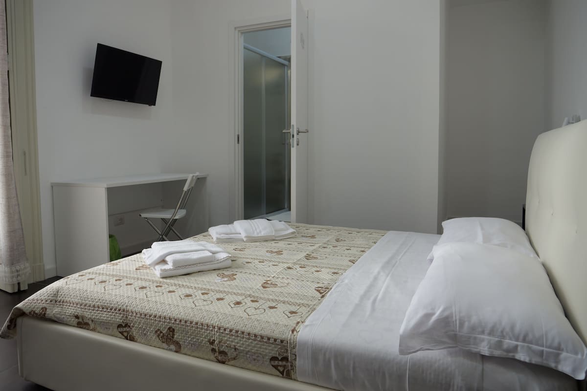 A bedroom with a bed, a TV mounted on the wall, a desk, and a door leading to a bathroom.