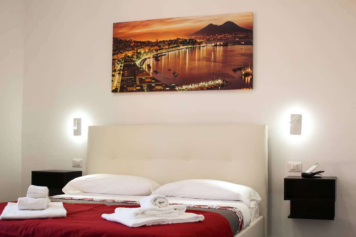 A hotel room with a bed, two bedside tables, a nightstand, and a wall-mounted painting of a cityscape.