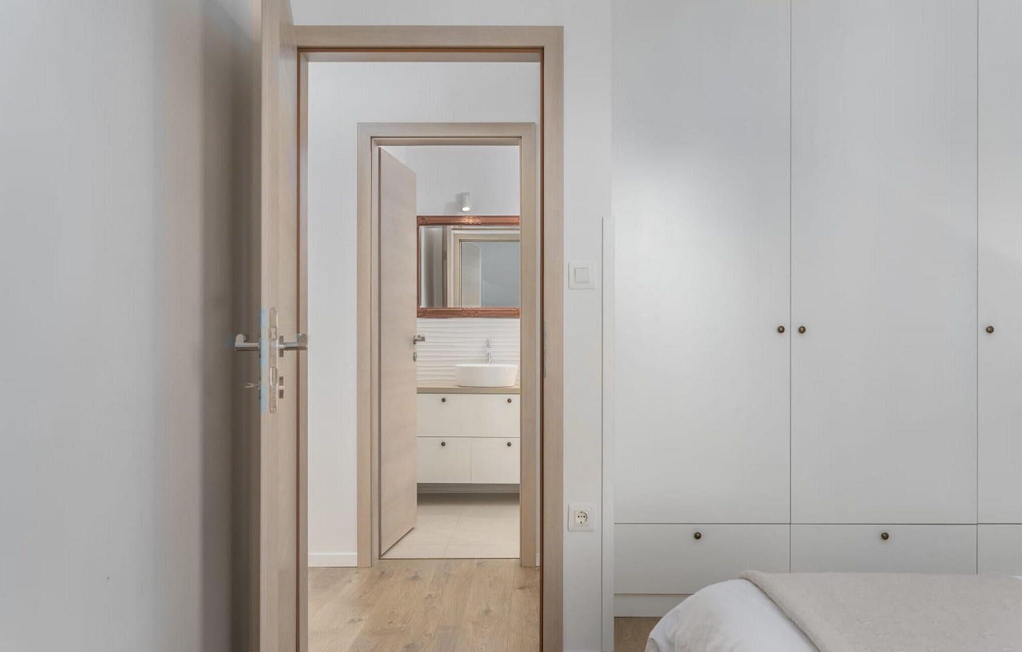 Un dormitorio con una puerta de madera que da a un baño. La habitación tiene una cama, un armario y un espejo sobre el lavabo.
