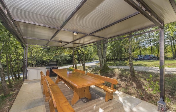 Une terrasse extérieure couverte, avec une table et des bancs en bois, un barbecue et une vue sur la forêt environnante.