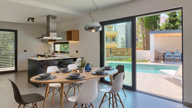 A modern kitchen with a central island, a dining area with a round table and chairs, and a view of a swimming pool through large glass doors.