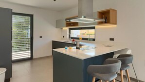 A modern kitchen with a central island, bar stools, a built-in oven, and a window with blinds.