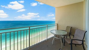 A balcony with a glass table and two chairs, overlooking a beach with clear turquoise water and a sandy shore. - Emerald Beach Resort 2235 (Panama City Beach)