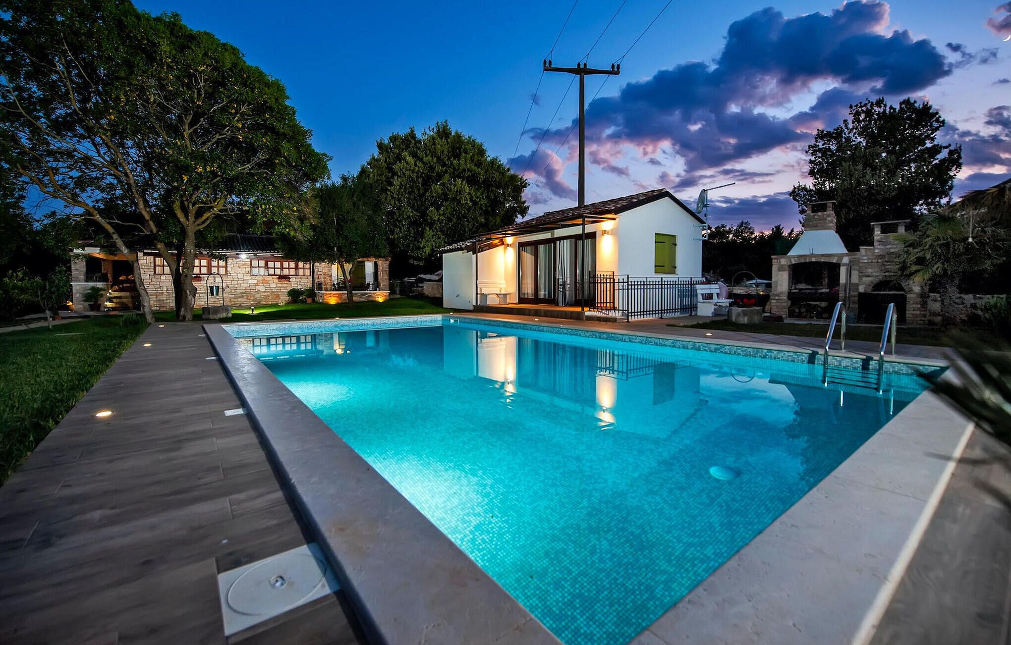 A swimming pool with a wooden deck, a stone house, and a barbecue area.