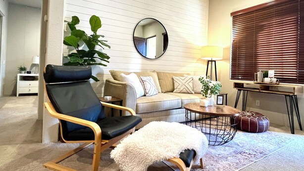 Un salon comprenant un canapé, un fauteuil, une table basse, une lampe, une table d’appoint, un miroir et une fenêtre avec des stores.