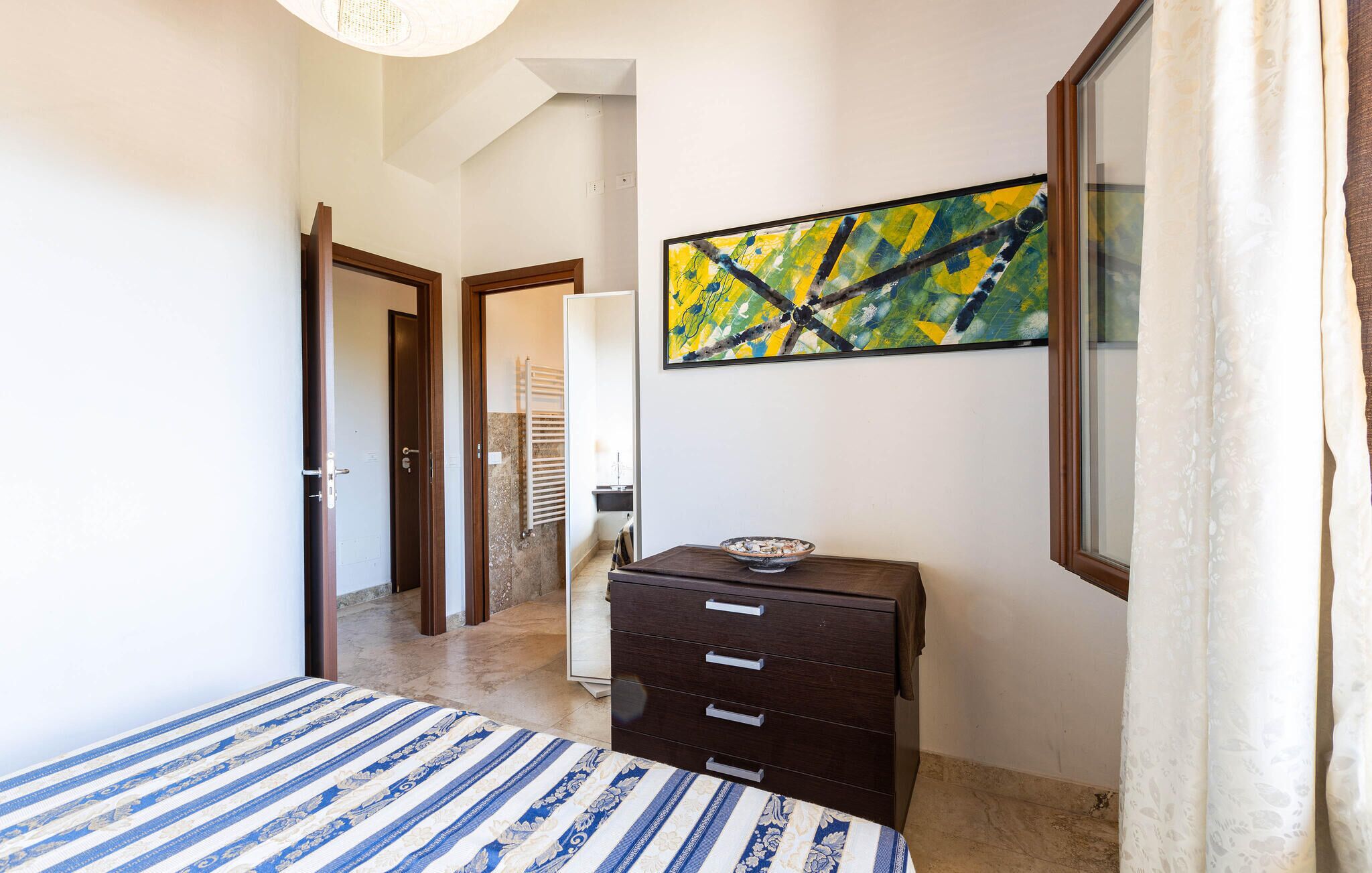 A bedroom with a bed, a dresser, a painting, and a bathroom visible through an open door.
