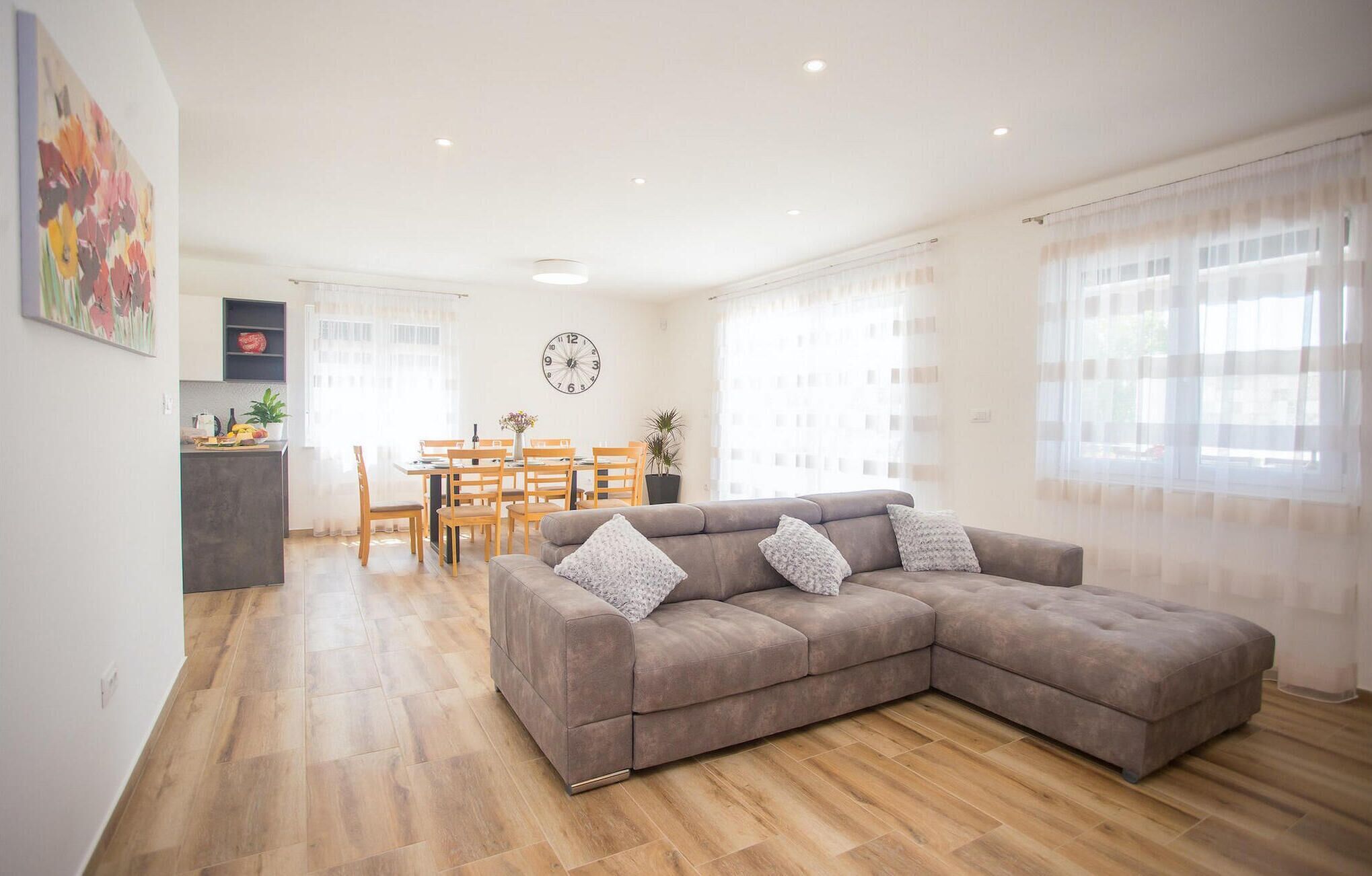 A modern living room with a sectional sofa, wooden flooring, a dining area with a table and chairs, and a kitchen area with a countertop and cabinets.