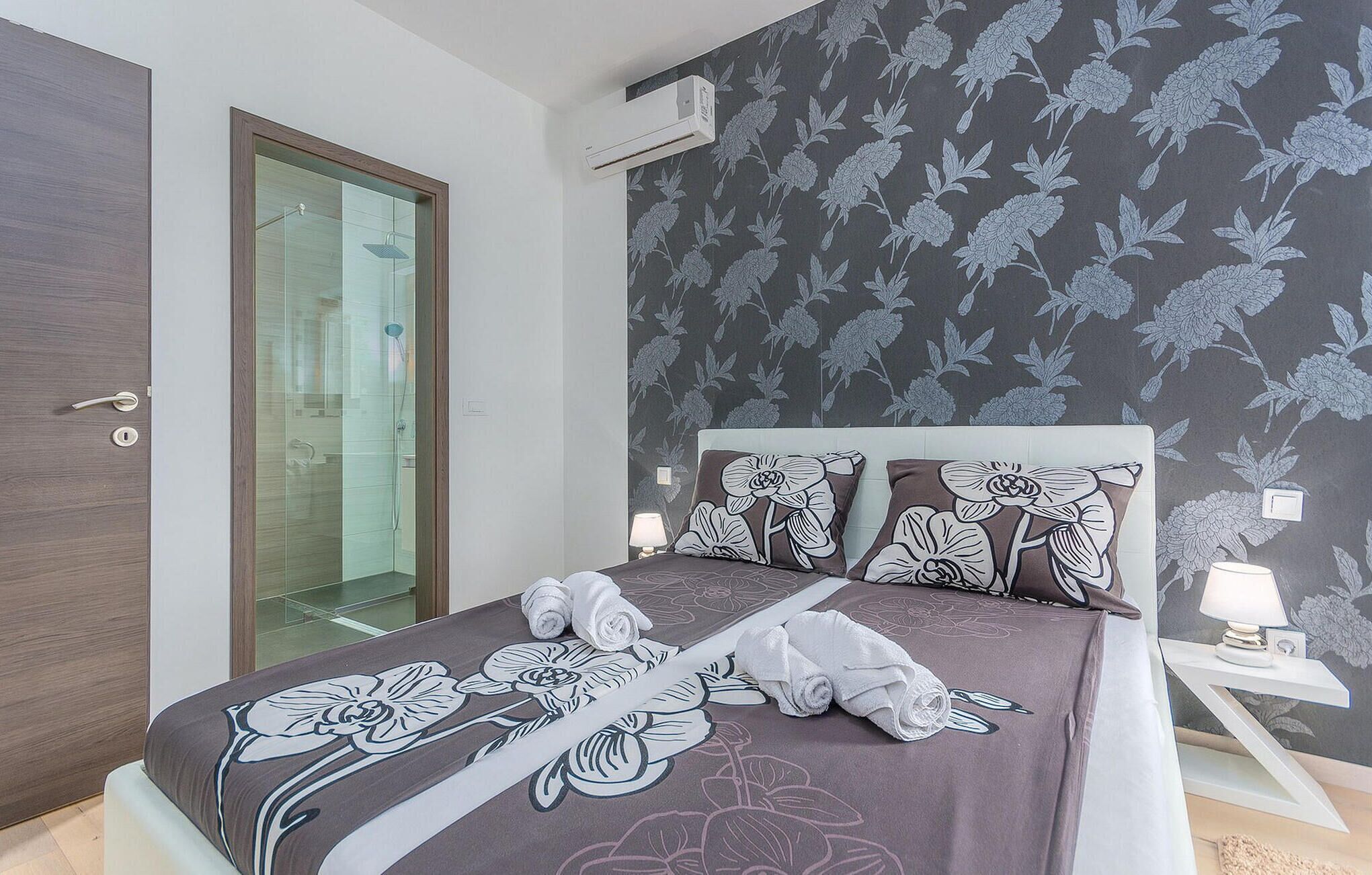 Une chambre à coucher avec un lit, une porte de douche en verre, une table de chevet avec une lampe et un papier peint à motifs floraux.