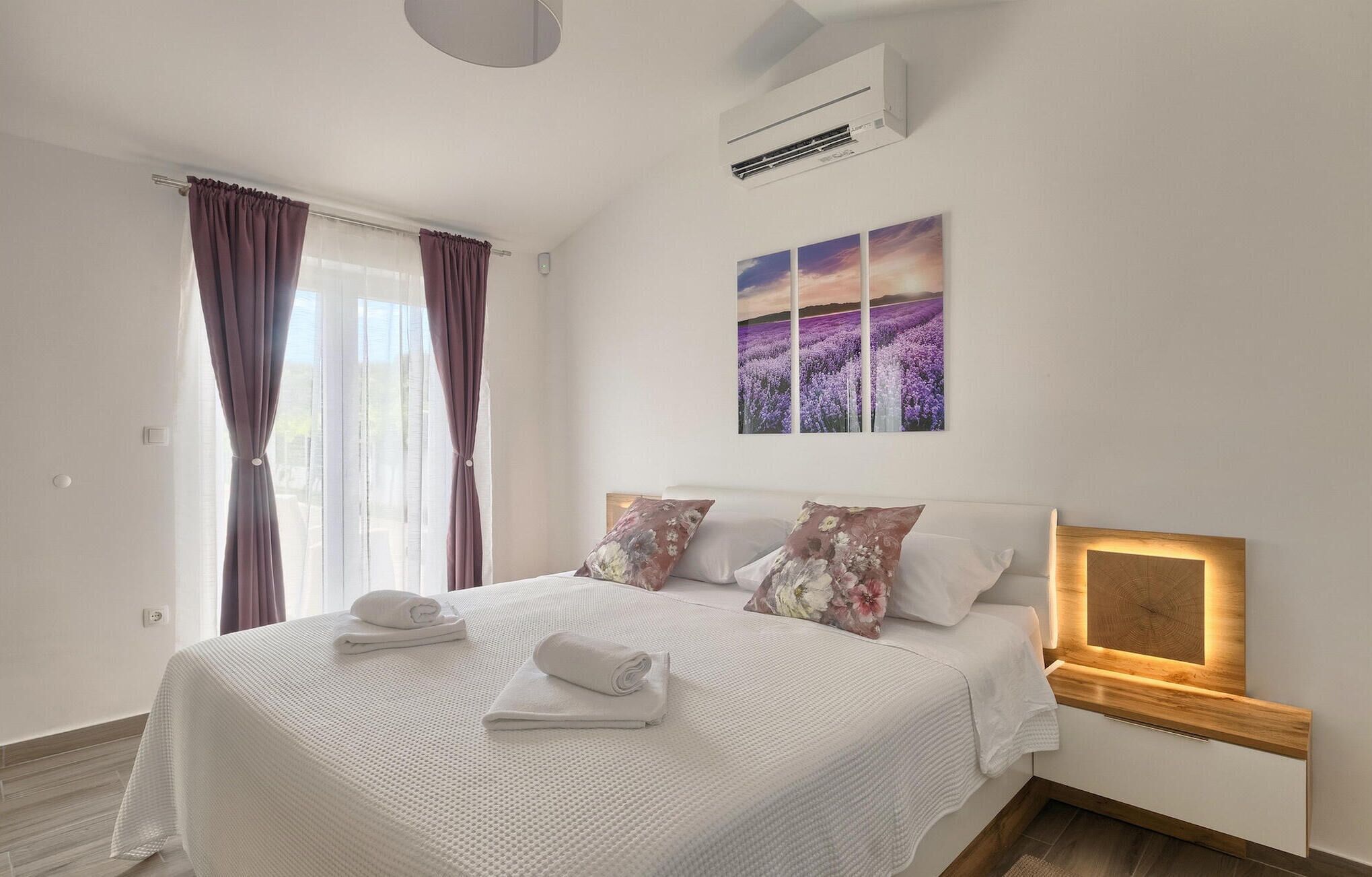 Une chambre dotée d’un grand lit, d’une tête de lit avec éclairage intégré, d’un climatiseur fixé au mur et d’une fenêtre avec des rideaux.