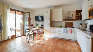 Une cuisine avec des meubles blancs, un sol carrelé, un coin repas avec des chaises et un téléviseur fixé au mur.
