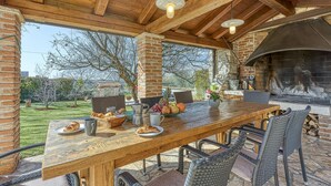 An outdoor patio with a wooden table set for dining, surrounded by wicker chairs, and a stone fireplace in the background.