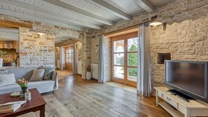 A spacious living room with a stone wall, wooden beams, a sofa, a coffee table, a TV, and a view through a glass door.