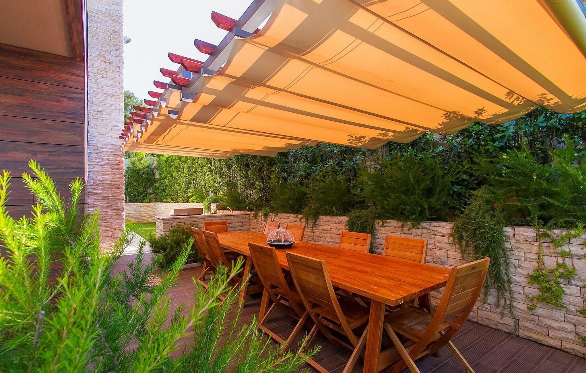 Een terras buiten met een houten tafel en stoelen, een pergola met een overkapping en een stenen muur met beplanting.