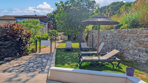 Un jardin avec un mur en pierre, un parasol en chaume et deux chaises longues.