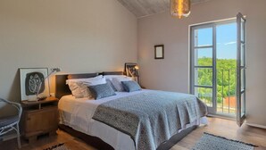 Un dormitorio con cama, mesitas de noche, una silla, un ventanal con vistas y una lámpara colgante.