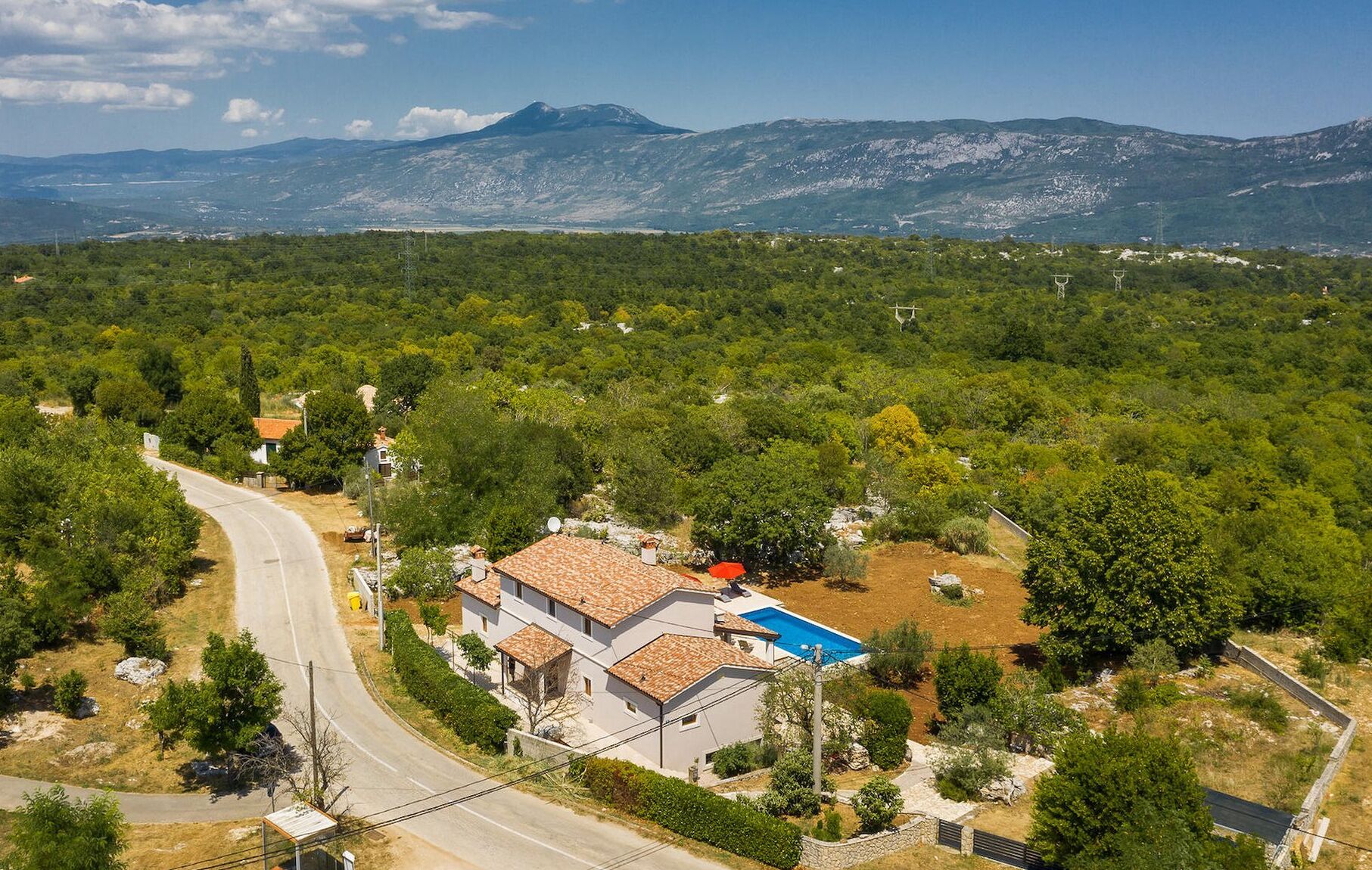 A residential area with a house featuring a tiled roof, a swimming pool, and a road curving through the landscape.
