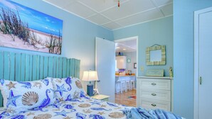 Une chambre à coucher avec un lit agrémenté d’une couvre-lit sur le thème de la mer, un tableau représentant une scène de plage et une vue sur la salle à manger adjacente.