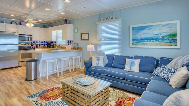Une cuisine moderne avec des meubles blancs, un coin bar et un canapé bleu dans un salon orné d’un tableau évoquant le bord de mer.