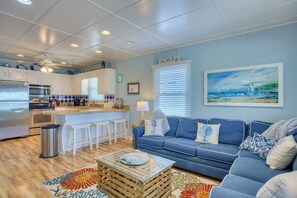 Een moderne keuken met witte kasten, een bar en een blauwe bank in een woonkamer met een schilderij van een kustlandschap.