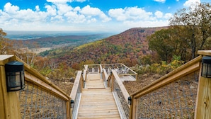 Eine Holztreppe führt zu einer Terrasse mit Stühlen, mit Blick auf ein bewaldetes Tal.