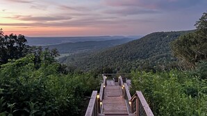 Eine Holzbrücke führt zu einer Aussichtsplattform mit Blick auf sanfte Hügel und einen Sonnenuntergang.