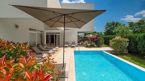 Un espace piscine extérieur moderne, doté de chaises longues et d’un grand parasol, entouré de verdure et surplombé d’un ciel bleu dégagé.
