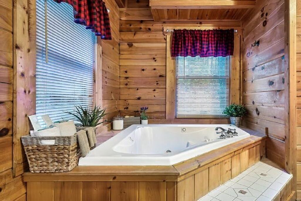 Une salle de bain rustique, aménagée dans une cabane en bois, avec une grande baignoire, une fenêtre avec des stores et un panier rempli de produits de toilette.