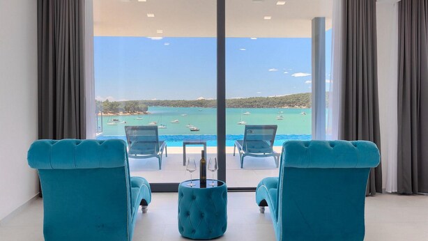 A room with a large sliding glass door leading to a balcony with a view of the sea and boats. There are two blue cushioned chairs and a small round table with a bottle and glasses on it. - Beautiful home in Medulin with sauna (Medulin)