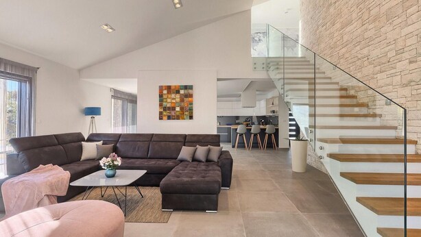 Un salon moderne avec un canapé d’angle, une table basse et un fauteuil rose. Un escalier avec des rampes en verre mène à un niveau supérieur.