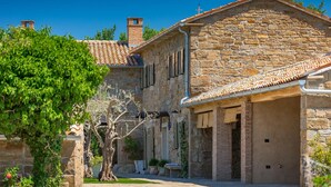 Un bâtiment en pierre avec un toit en tuiles, un porche abrité et un jardin agrémenté d’arbres et d’arbustes.
