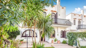 A courtyard with a white building featuring a balcony with wrought iron railings, a palm tree, and a small garden area with a white chair and potted plants.