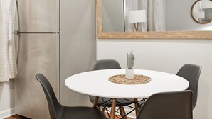 Un espacio habitable compacto con una mesa de comedor redonda blanca, cuatro sillas, una pequeña planta en maceta, un espejo grande y un refrigerador blanco.