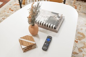Une table blanche sur laquelle se trouvent un livre intitulé « Design for Living », une télécommande et un vase décoratif contenant des fleurs séchées.