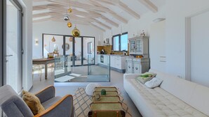 Une cuisine moderne avec des meubles blancs, un coin repas doté de tables en verre et un salon avec un canapé blanc et un pouf à motifs.