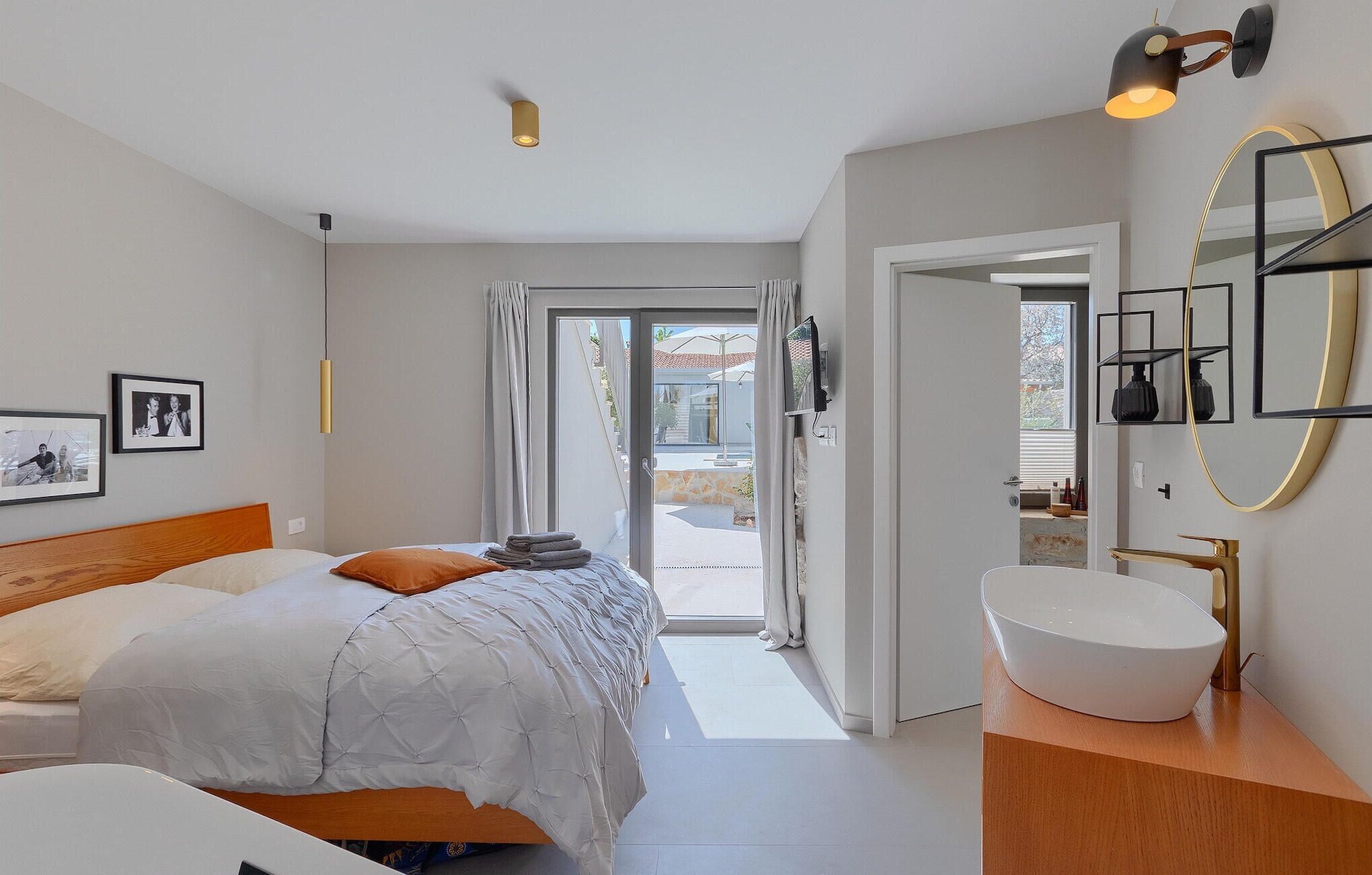 A modern bedroom with a bed, a large window, a bathroom with a sink and mirror, and a hallway leading to an outdoor area.