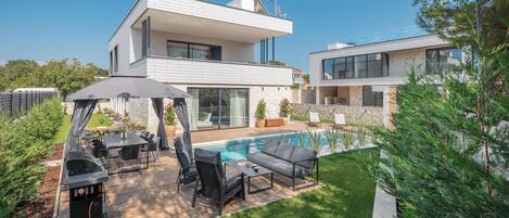 Ein modernes, zweistöckiges Haus mit Schwimmbad, Sitzgelegenheiten im Freien und einer Pergola.