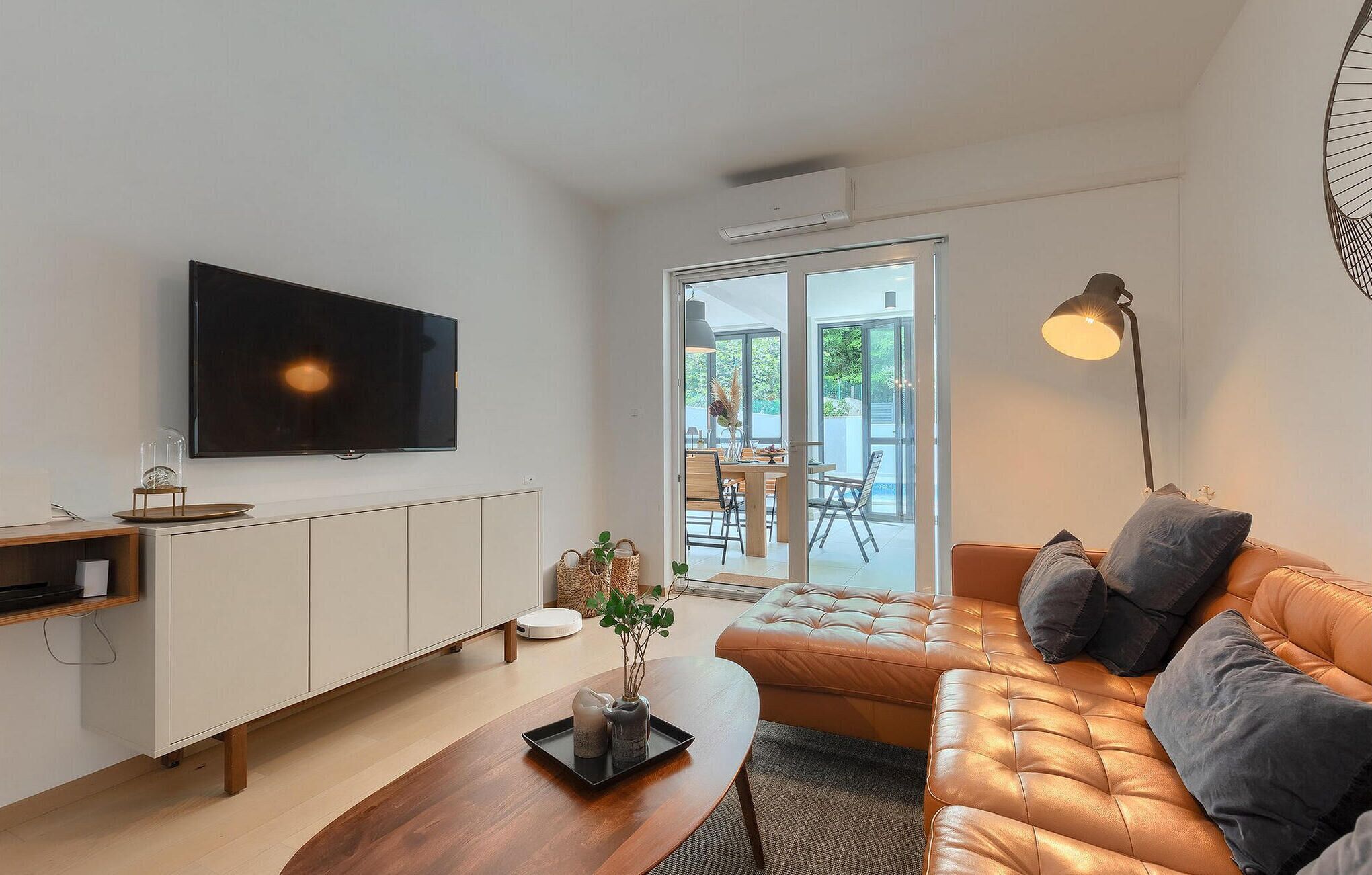 A modern living room with a flat-screen TV, a wooden coffee table, a leather sofa, and a dining area visible through glass doors.