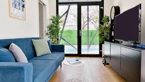 Una sala moderna con un sofá azul, una mesa de centro redonda, un televisor de pantalla plana sobre un mueble bajo, y vistas a un seto verde y un edificio a través de una puerta de cristal.