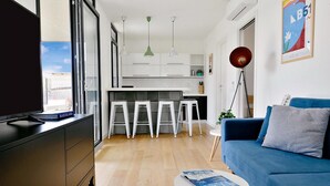 Una sala de estar moderna con televisor, un sofá azul y una zona de cocina con armarios blancos y taburetes de bar.