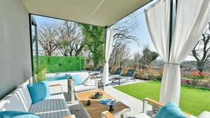 Un área de estar al aire libre moderna con sofás blancos y cojines azules, una mesa de madera y vista a un césped verde y árboles.