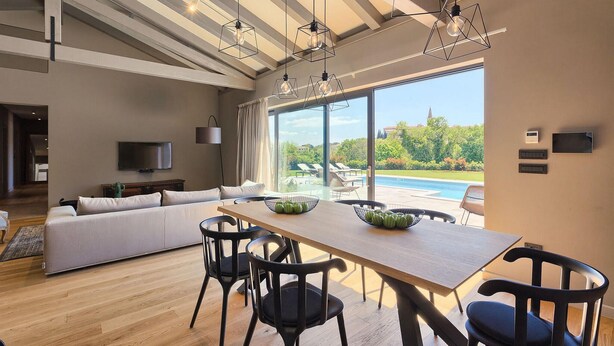 Ein modernes Wohnzimmer mit einem Holzesstisch, schwarzen Stühlen, einer großen Couch und einem Fernseher. Durch große Fenster ist ein Pool sichtbar.