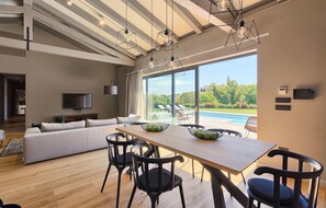 Un salon moderne avec une table à manger en bois, des chaises noires, un grand canapé et un téléviseur. On peut apercevoir une piscine à travers de grandes fenêtres.