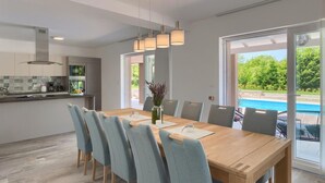 A modern dining area with a wooden table and grey upholstered chairs, a kitchen with patterned backsplash, and a view of a swimming pool through large sliding doors. - Awesome home in Pazin with sauna (Pazin)