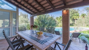 A covered patio with a table set for dining, surrounded by chairs, and overlooking a swimming pool.