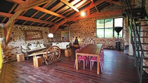 Una stanza rustica con parete in pietra, travi in legno, una panca di legno, un grande tavolo di legno con sedie rosa e una ruota di carro in legno, oltre a una scala.