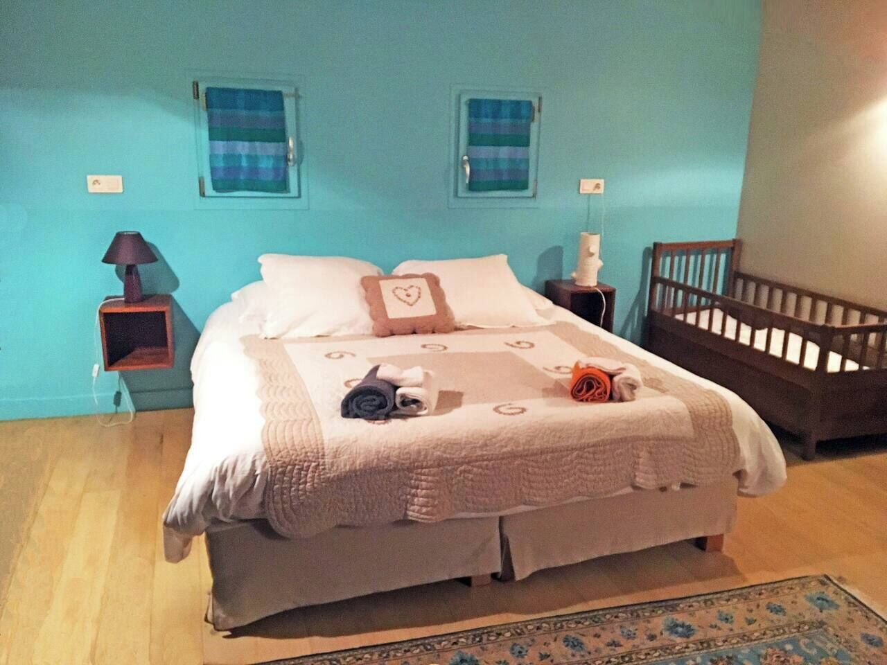 Een net opgemaakt bed met een witte dekbedovertrek en kussens, een klein nachtkastje met een lamp, een houten babybedje en een blauwe muur met twee ramen.