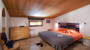 A bedroom with a sloped ceiling, a bed with an orange bedspread, a dresser, a chair, and a small table with a lamp. - Au calme & au coeur de la station | 50m remont ées (Les Deux Alpes)
