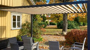 Une terrasse couverte avec des fauteuils en osier, une pergola et un jardin aux couleurs automnales.