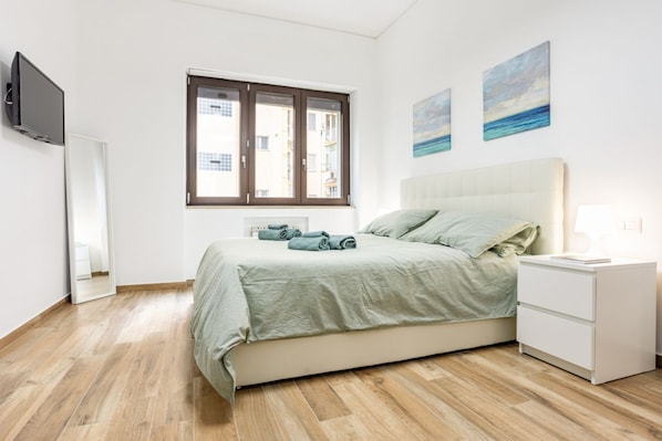 A bedroom with a bed, a nightstand, a TV mounted on the wall, and a window with a view of buildings.